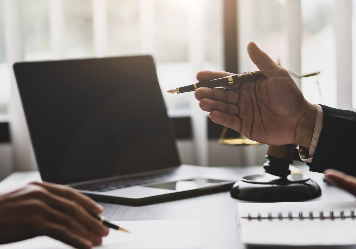 Two people in a meeting with a laptop and documents on a table, one holds a pen gesturing, while the other listens.